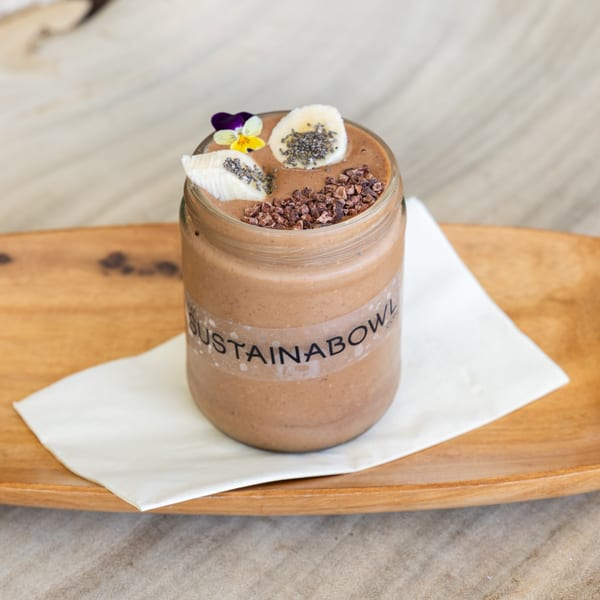 Sustainabowl chocolate smoothie with banana, chia seeds, cacao nibs, and a flower on a wooden tray.