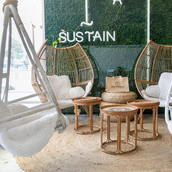 Rattan egg chairs with white cushions, small round tables, and a "Sustainabowl" bag on a jute rug.