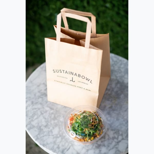 Sustainabowl branded paper bag and a healthy poke bowl on a marble table outdoors.