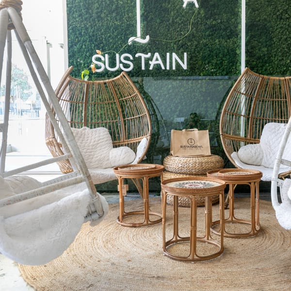 Cozy seating area with wicker chairs, small tables, and a "Sustain" sign on a moss wall.