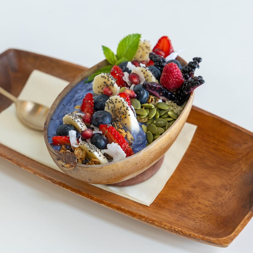 A vibrant blue smoothie bowl in a coconut shell, topped with fresh berries, banana, seeds, and mint.