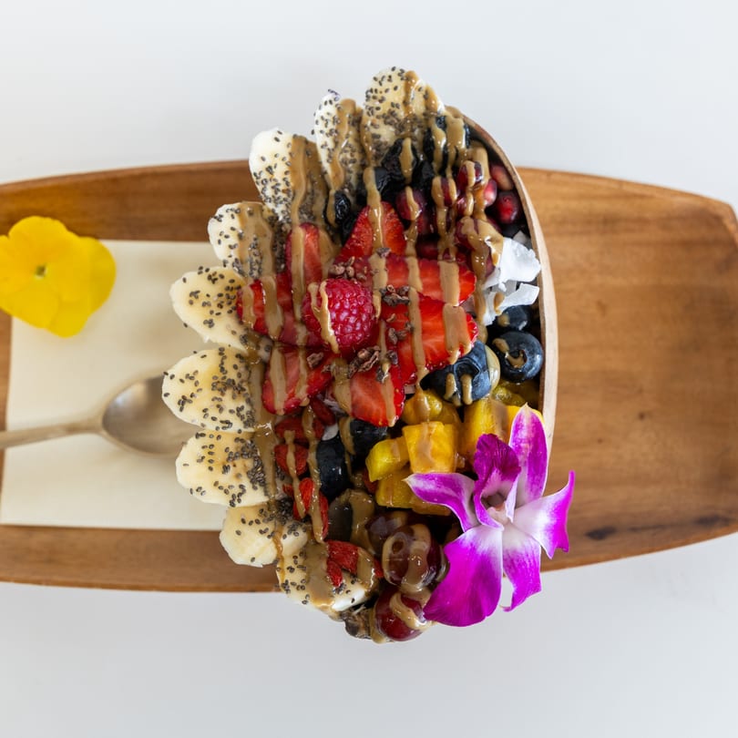 Colorful fruit bowl with bananas, berries, mango, nuts, and a purple orchid on a wooden tray.