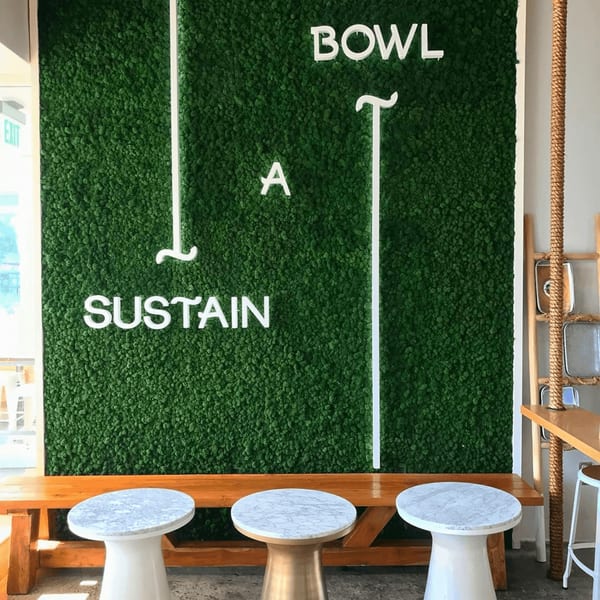 A green moss wall with "SUSTAIN A BOWL" in white letters, above a wooden bench and three small tables.