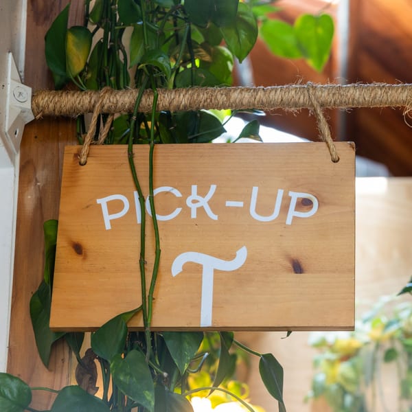 Wooden "PICK-UP" sign with white letters, hanging by rope, surrounded by green plants.