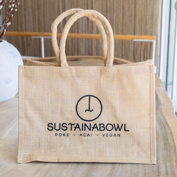 Sustainabowl branded reusable jute tote bag with black logo and text on a wooden table.