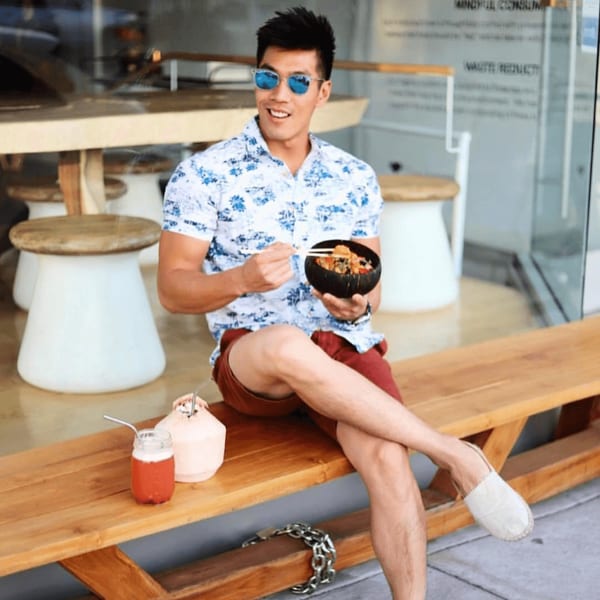 A man in sunglasses and a floral shirt eats a Sustainabowl dish on a wooden bench.