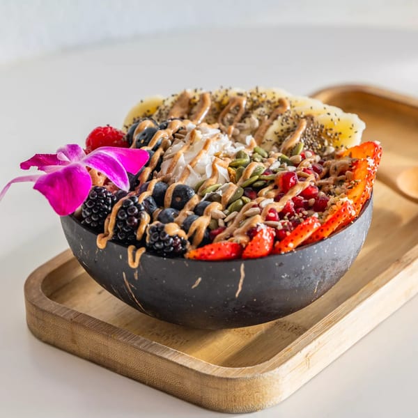 A vibrant Sustainabowl smoothie bowl in a coconut shell, topped with fruit, nuts, and a pink orchid.