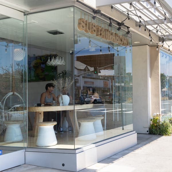 A woman sits at a wooden table inside a glass-fronted cafe with "SUSTAINABLE" signage.
