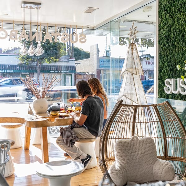 Two people eating healthy food at a wooden table inside Sustainabowl, with large windows overlooking a street.