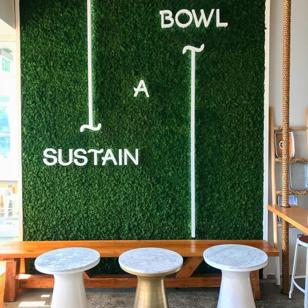 A green moss wall with white neon letters spelling "BOWL A SUSTAIN" above a wooden bench and round tables.