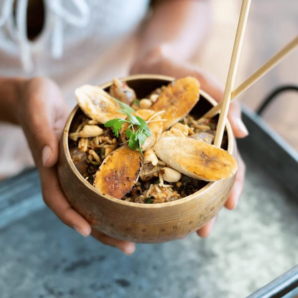 Hands holding a Sustainabowl with plantains, nuts, and cilantro, with chopsticks.