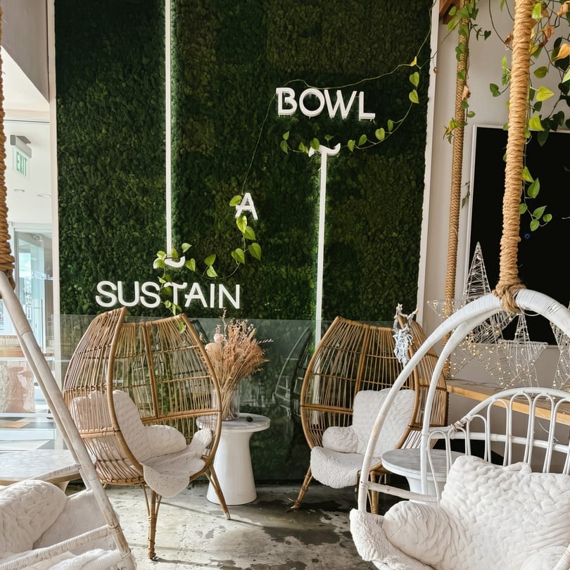 Cozy restaurant interior with hanging chairs, a moss wall, and neon signs that read "SUSTAIN A BOWL."