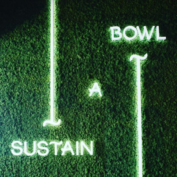 Neon signs spelling "BOWL A SUSTAIN" on a vibrant green moss wall.