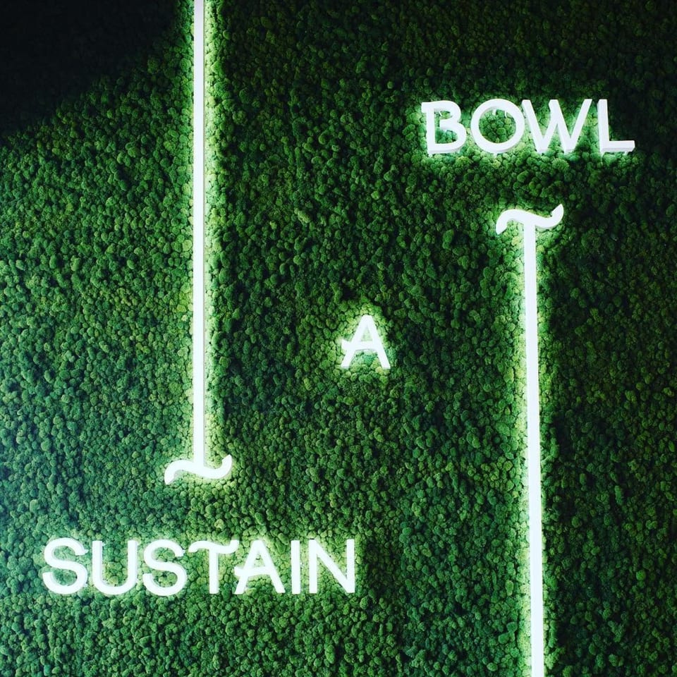 Neon signs spelling "BOWL A SUSTAIN" on a vibrant green moss wall.