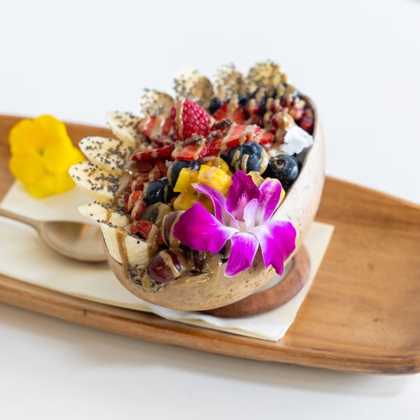 A vibrant fruit bowl with bananas, berries, mango, and nuts, drizzled with sauce and garnished with an orchid.