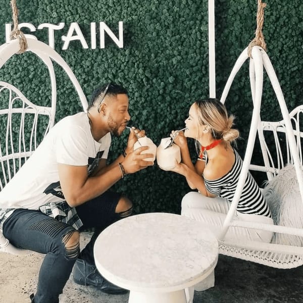 A couple in white hanging chairs sips from coconuts against a green moss wall with "SUSTAIN" lettering.