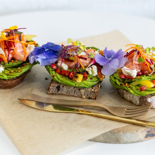 Three avocado toasts with salmon, edible flowers, and microgreens on a wooden board with gold cutlery.