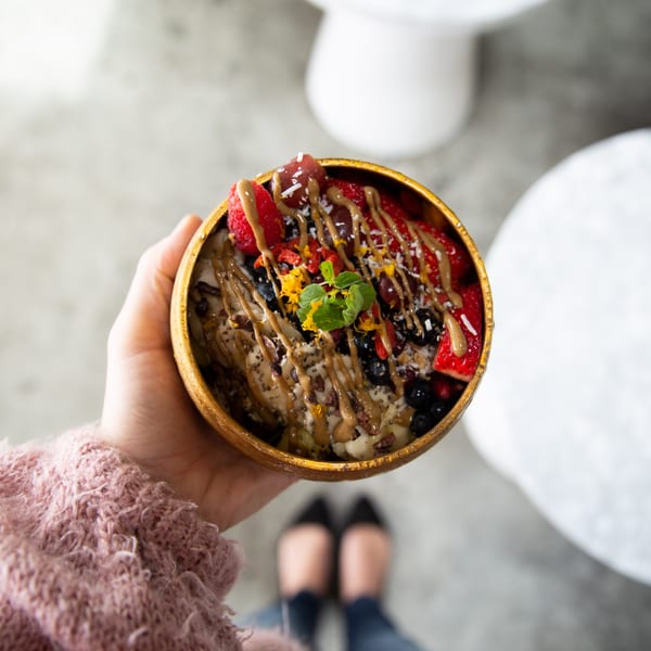 A hand holds a Sustainabowl with strawberries, blueberries, nuts, and a drizzle of sauce.