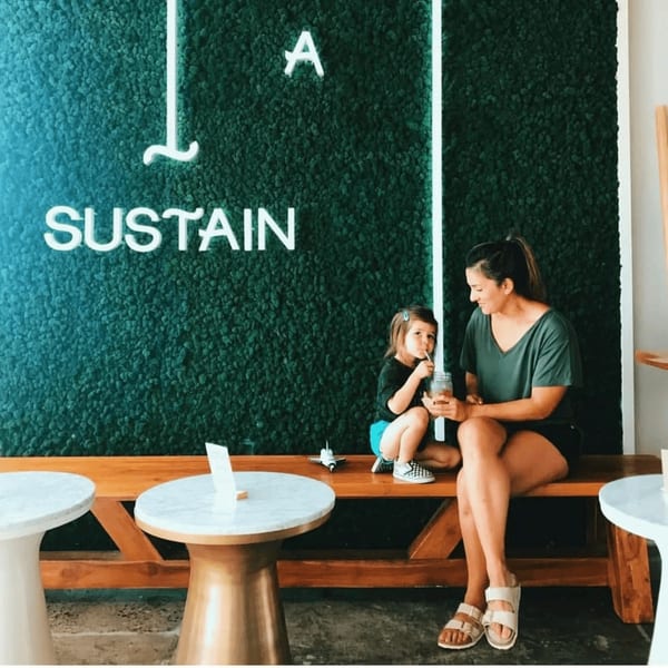 A woman and child sit on a wooden bench in front of a green moss wall with a neon "Sustain" sign.