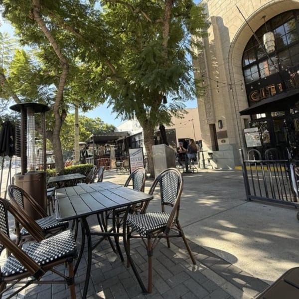 Outdoor patio seating with black tables and chairs, surrounded by trees and a building facade.