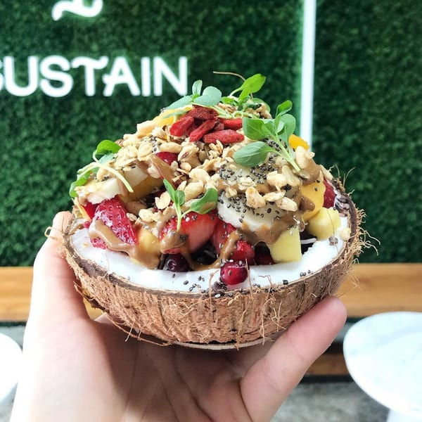 A hand holds a coconut bowl filled with fruit, granola, chia seeds, and a drizzle of sauce.