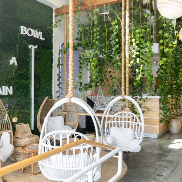 Restaurant interior with hanging white wicker chairs, lush green plants, and a moss wall with "BOWL A" signage.